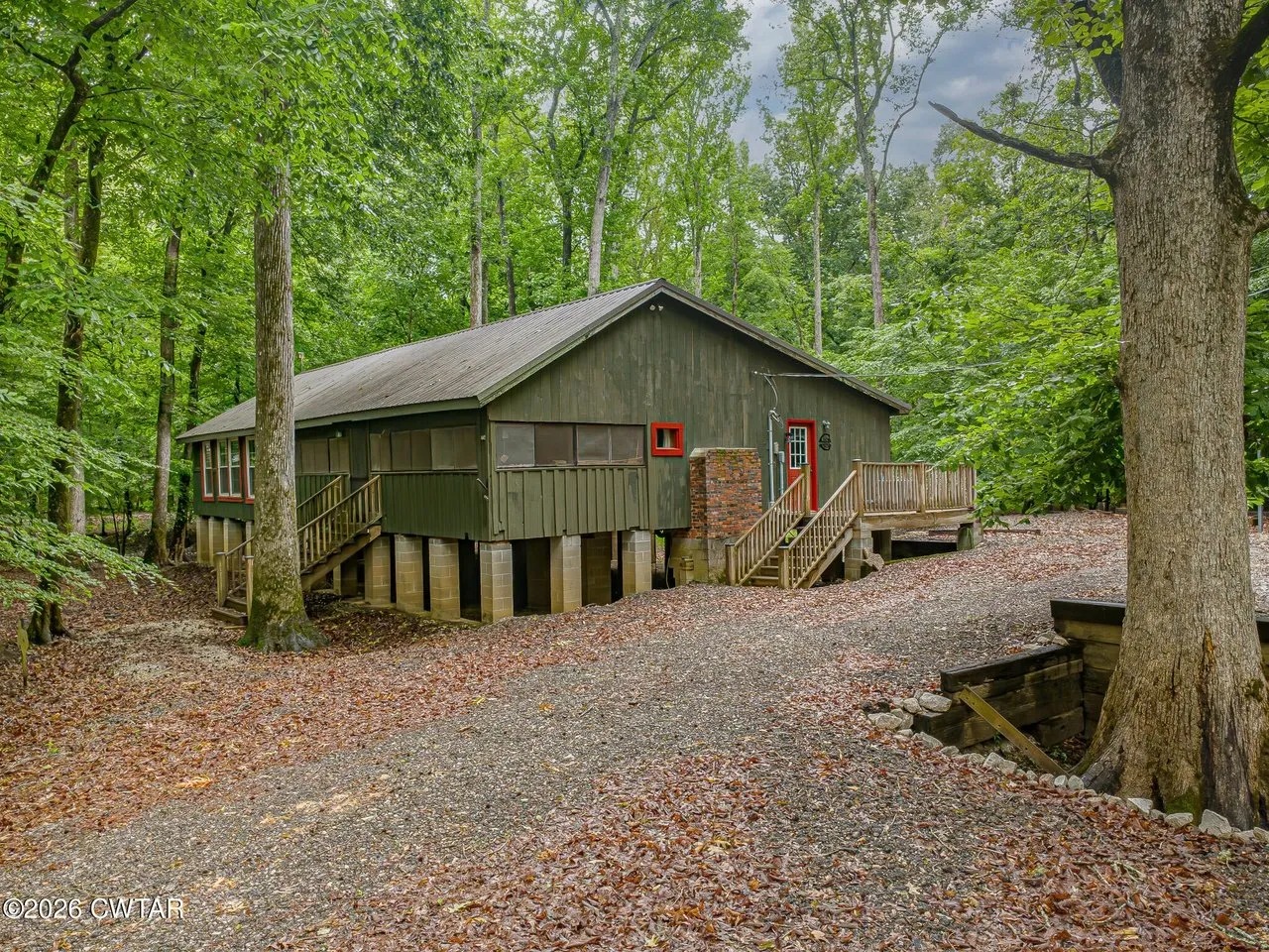 The cabin from the gravel drive, green siding raised on piers under mature hardwoods