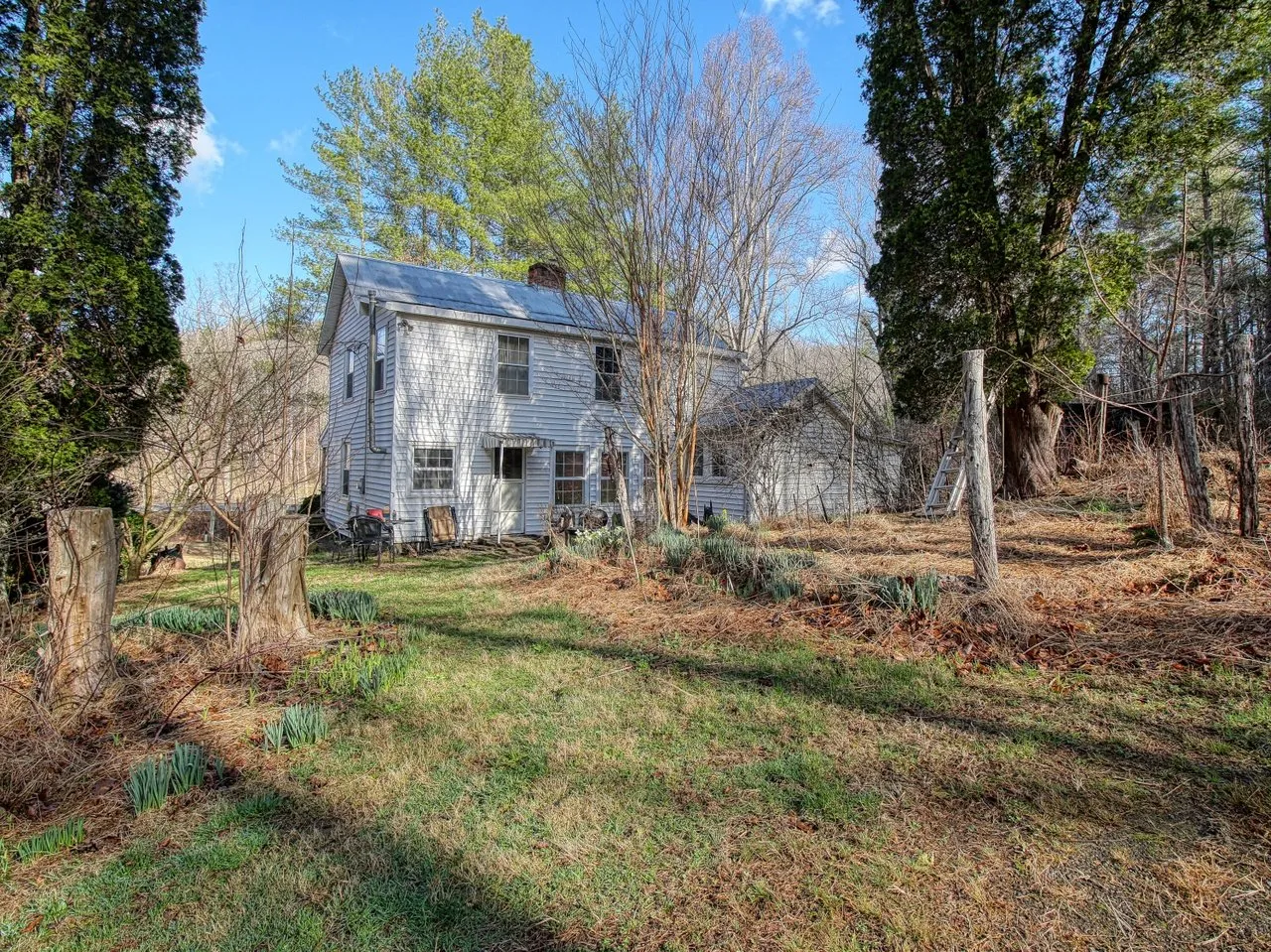 The farmhouse from the yard, white siding and a metal roof under mature trees