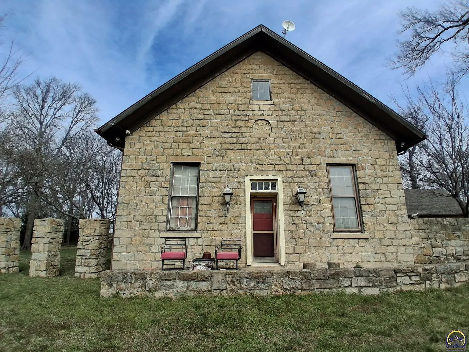 The stone schoolhouse from the front with its red door, the date stone in the gable and two chairs on the step