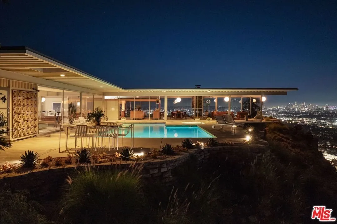 The house from the drive at night, pool glowing and the city beyond the edge