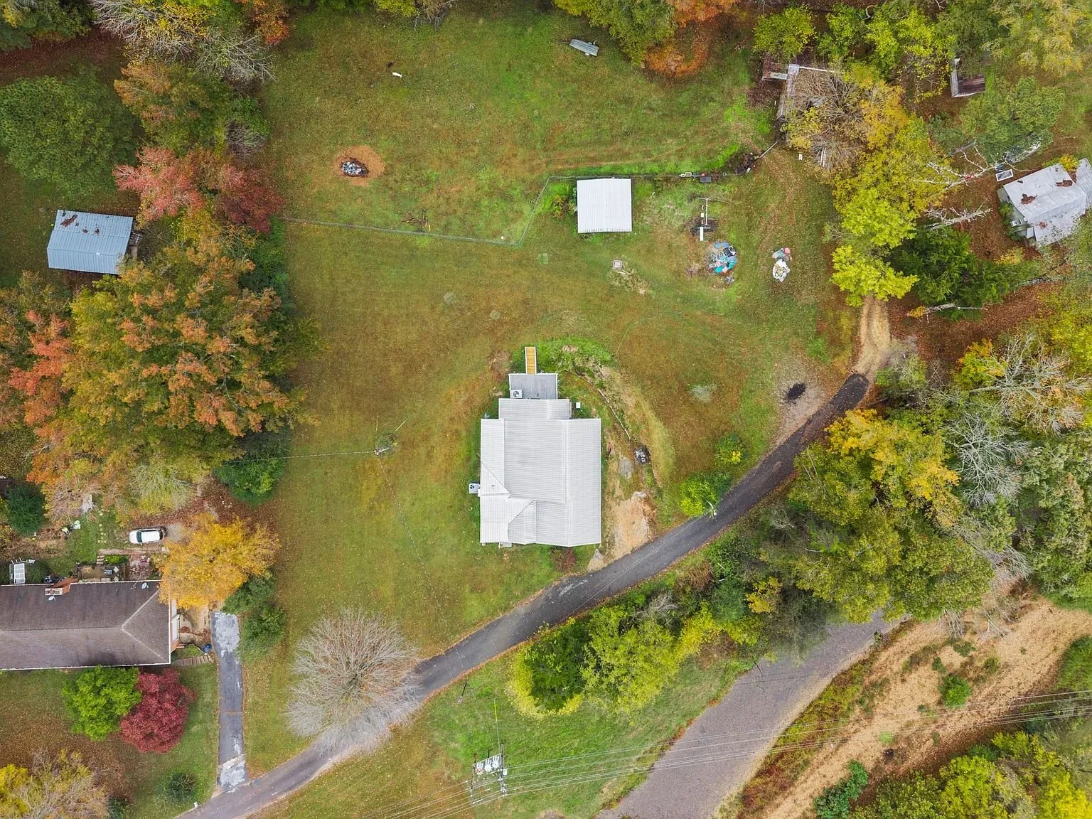 Top-down aerial of the lot: the house and its metal roof, the loop drive, and outbuildings toward the tree line