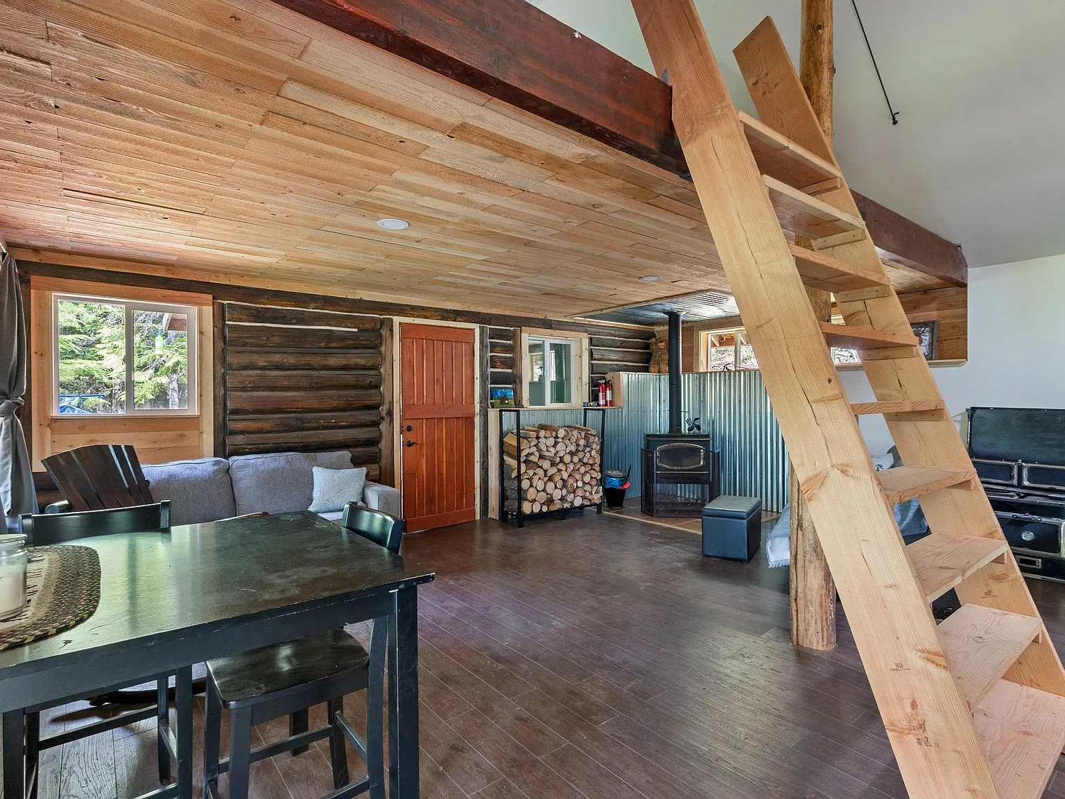 The open living area with a wood stove, gray sofa and log-paneled ceiling