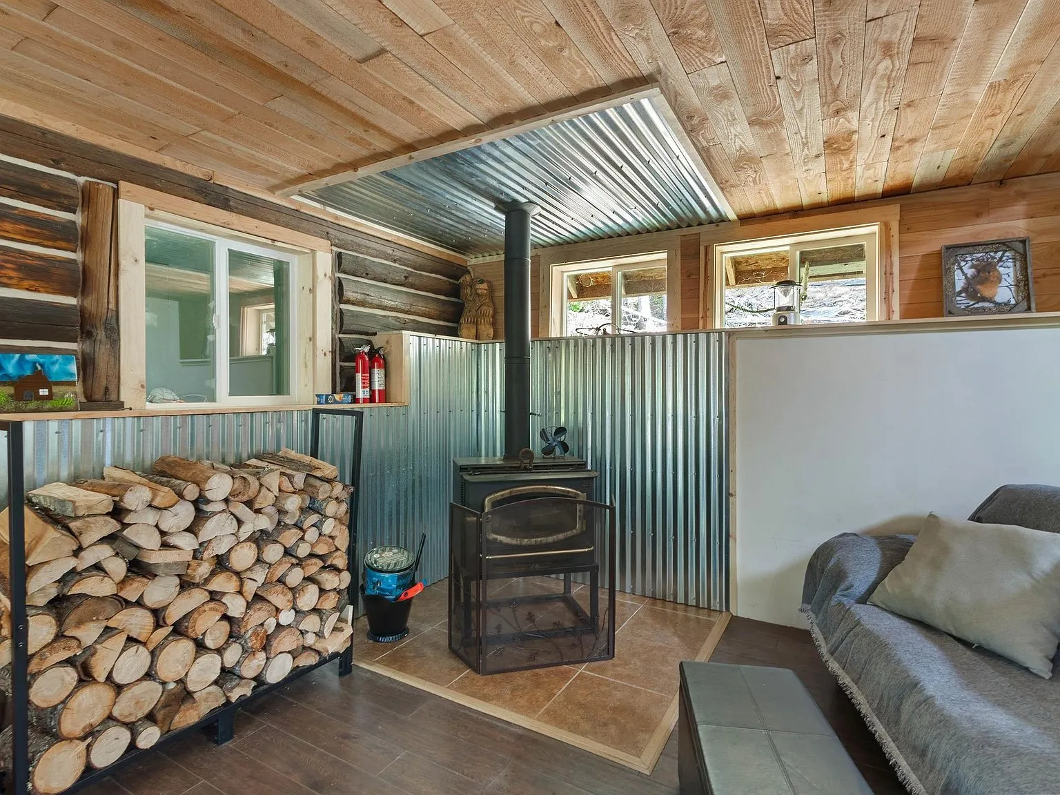 A second wood stove view with firewood storage and a sofa along the metal-paneled wall