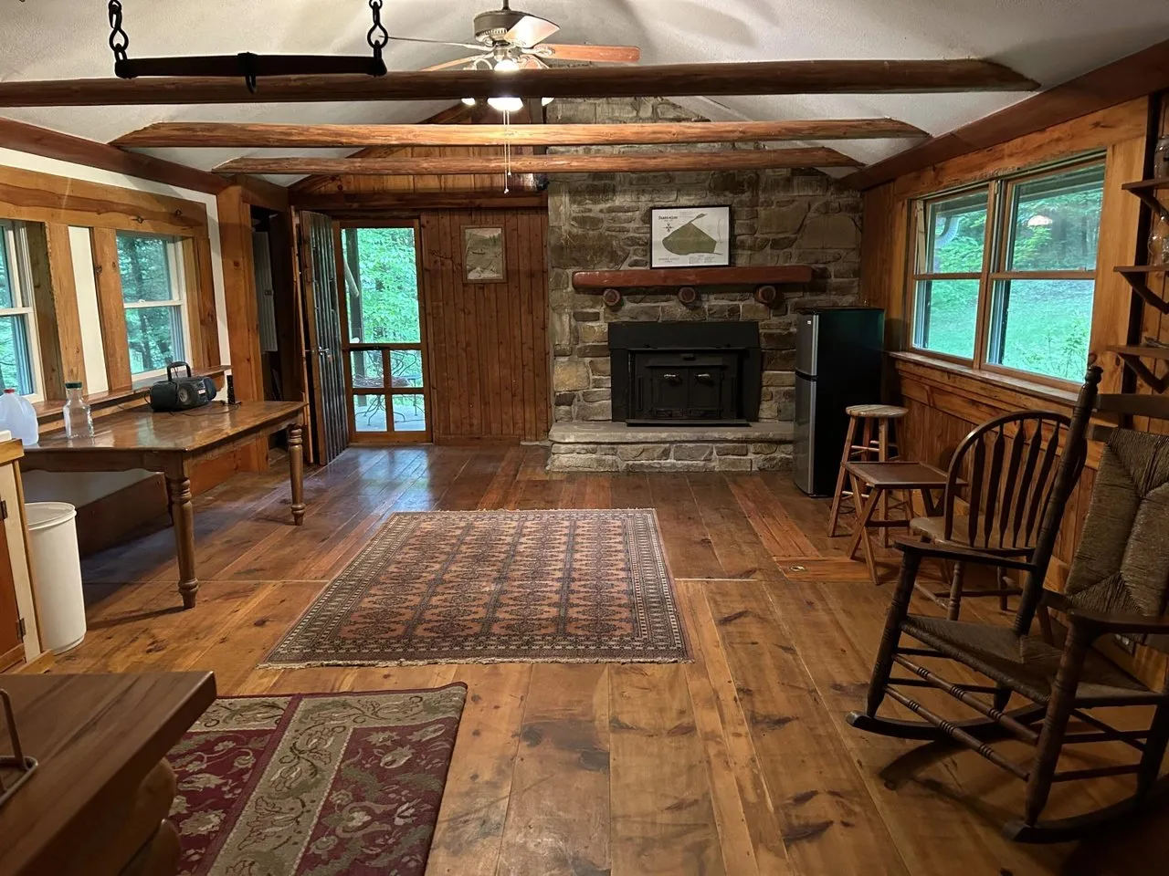 Main room of the spruce cabin: stone fireplace, exposed beams, wood floors and a wall of windows