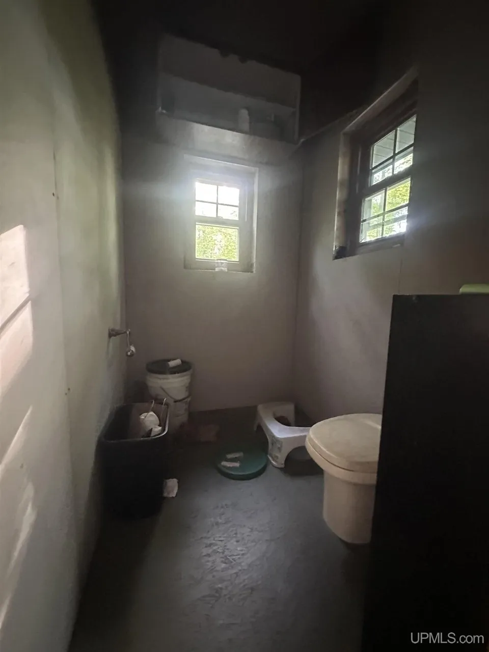 A small unfinished room with a bucket toilet and pails under the window