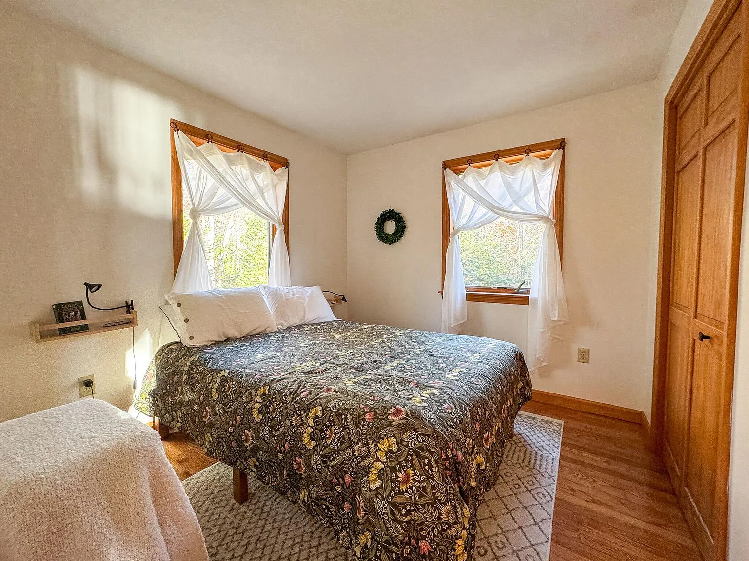 A bedroom with a patterned quilt, curtained windows and wood trim