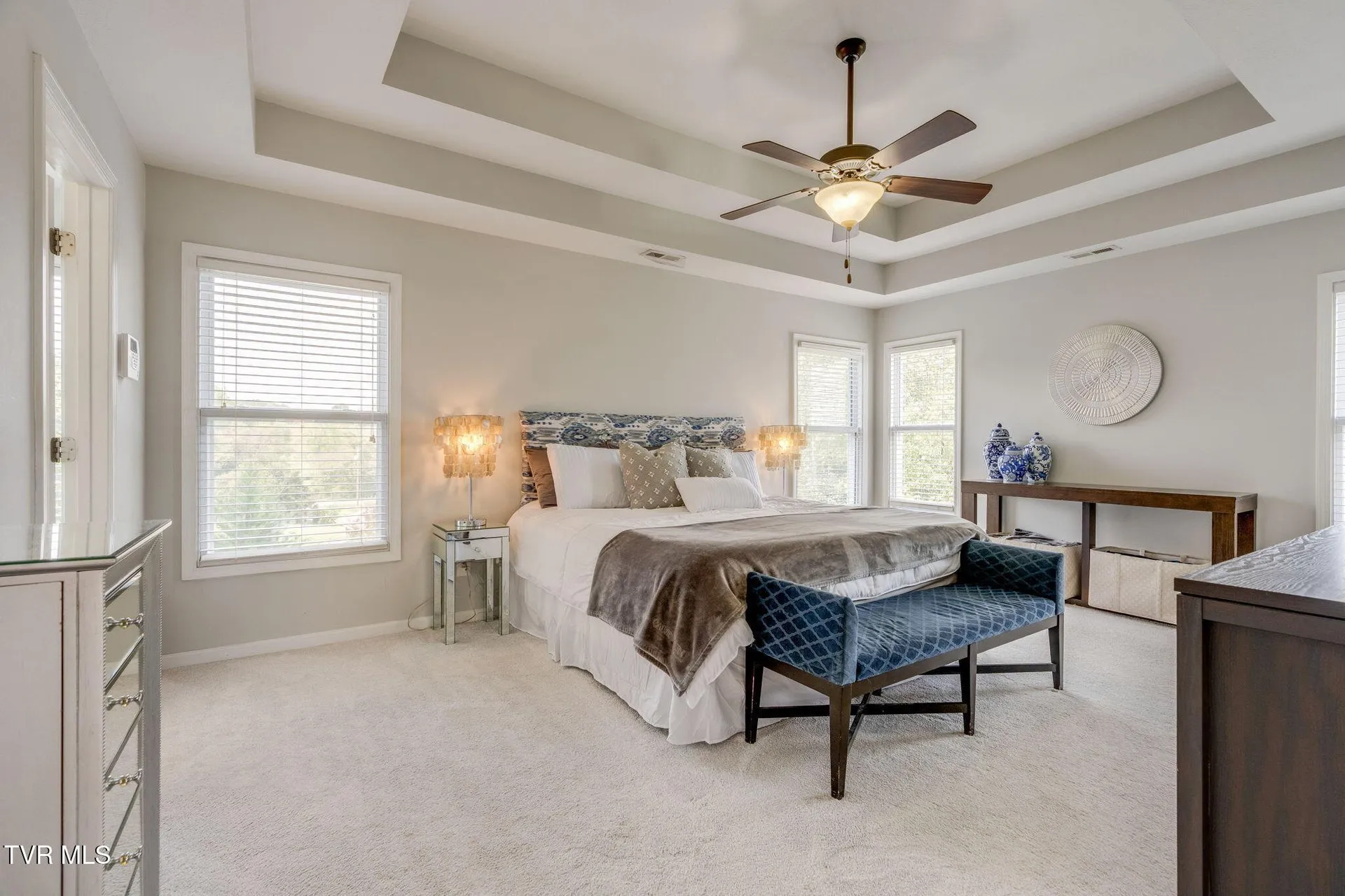 The primary bedroom with its tray ceiling, ceiling fan and windows on two walls