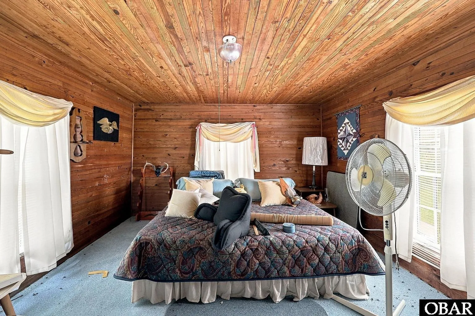 The wood-paneled bedroom with a bed, box fan and curtains
