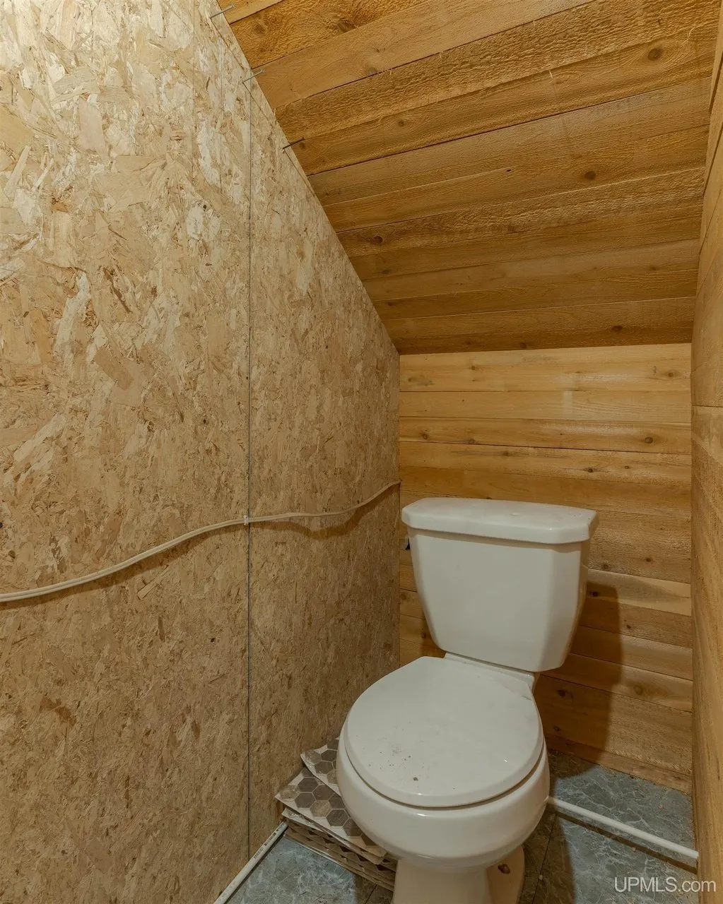 A toilet in a small room under the stairs, bare board walls