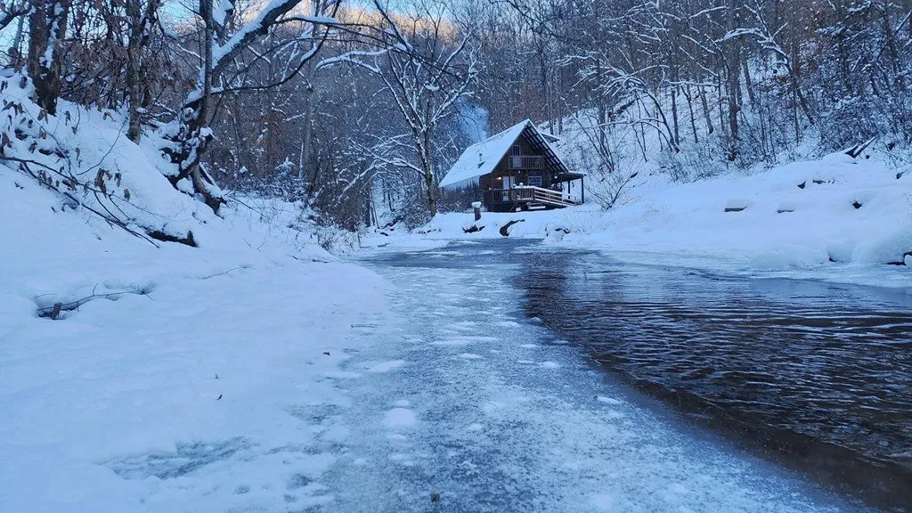 The creek frozen at the edges with the cabin behind it