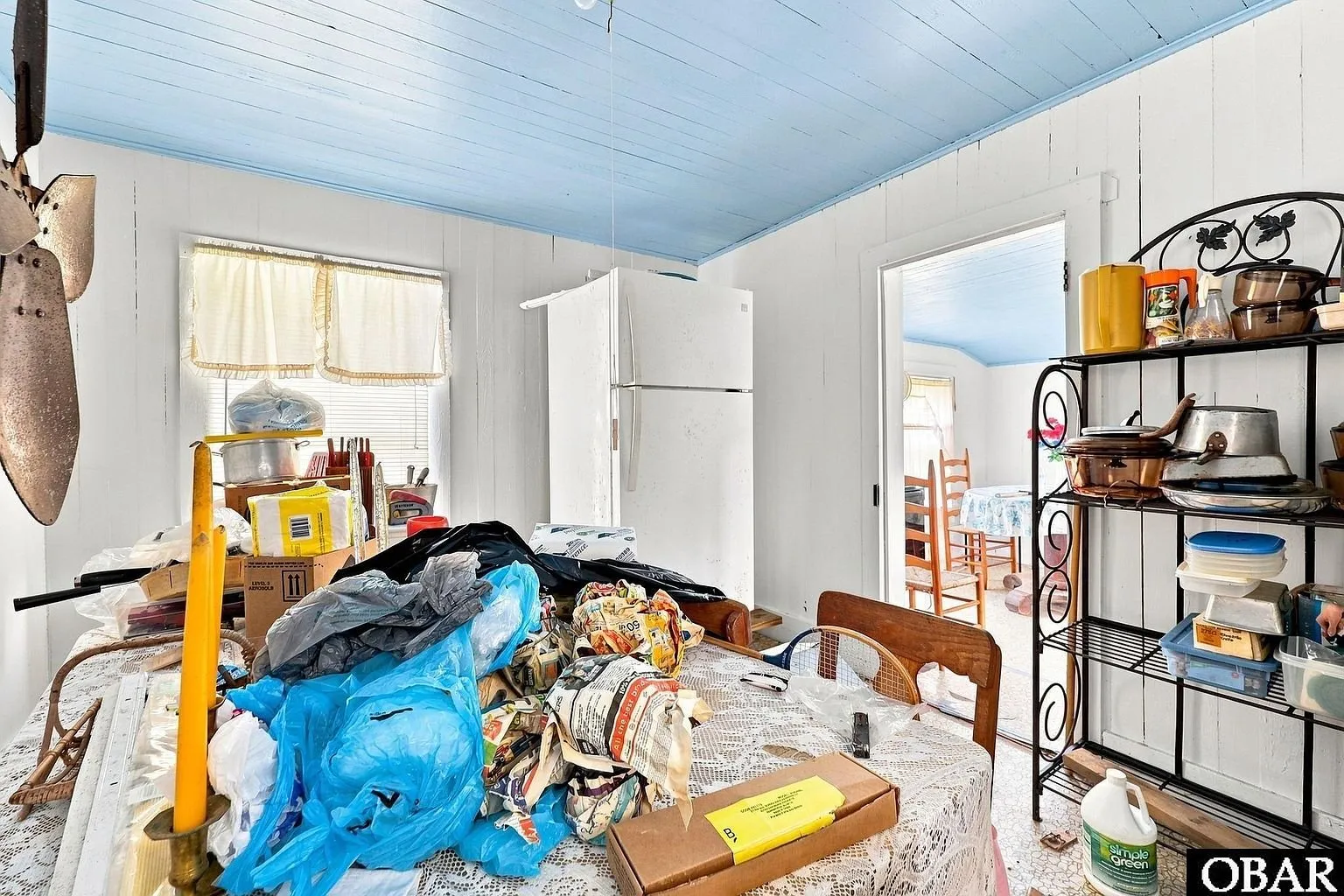 The kitchen with the refrigerator, shelving and a table covered in belongings