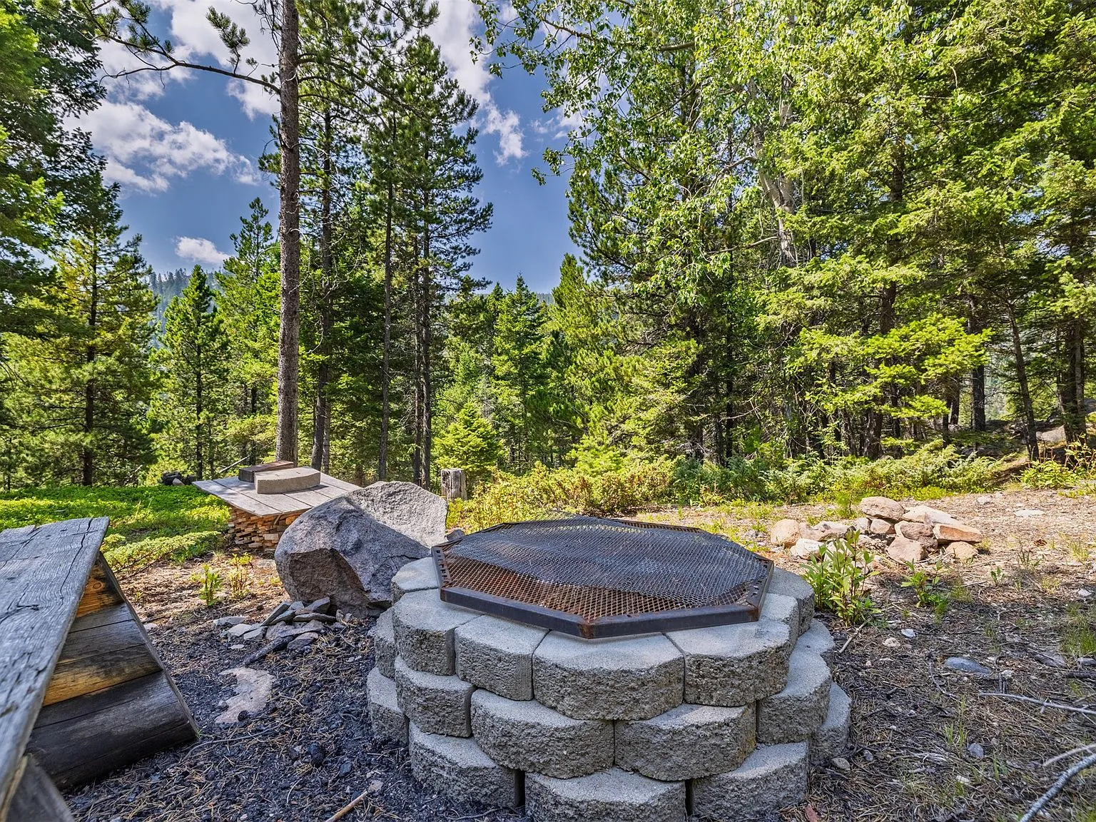 The stone fire pit with a metal grate cover, set among boulders and pines