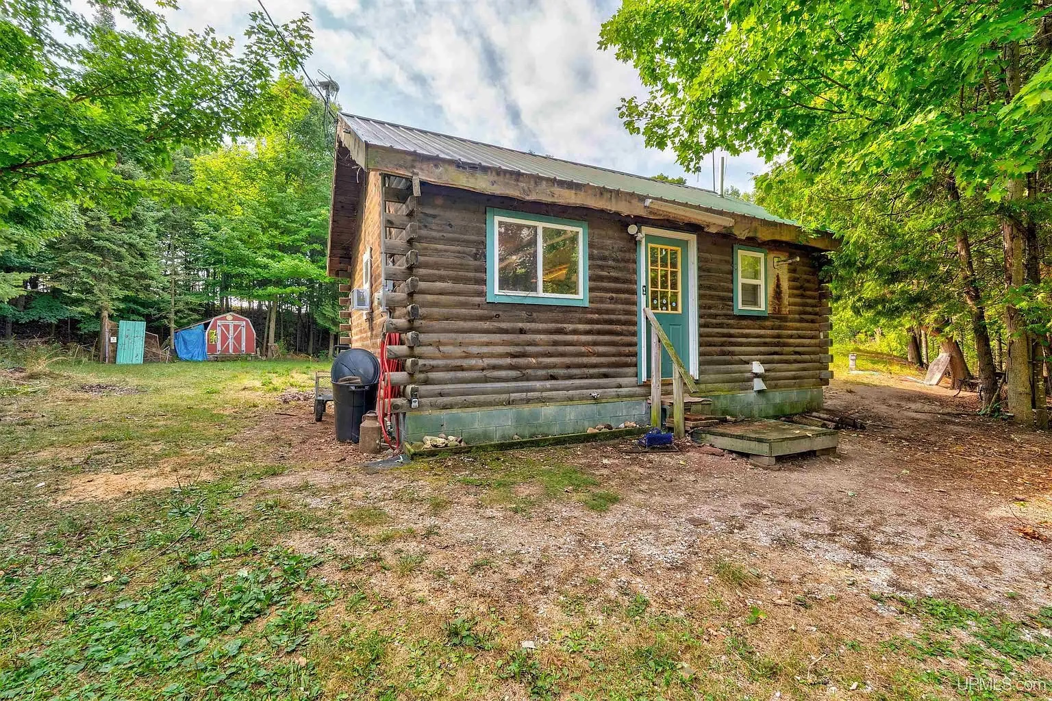 The round-log cottage with blue trim, a propane tank and the well head beside it