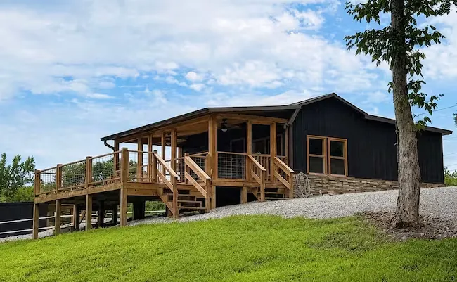 The barn-style tiny home with a wraparound deck, seen across the open yard