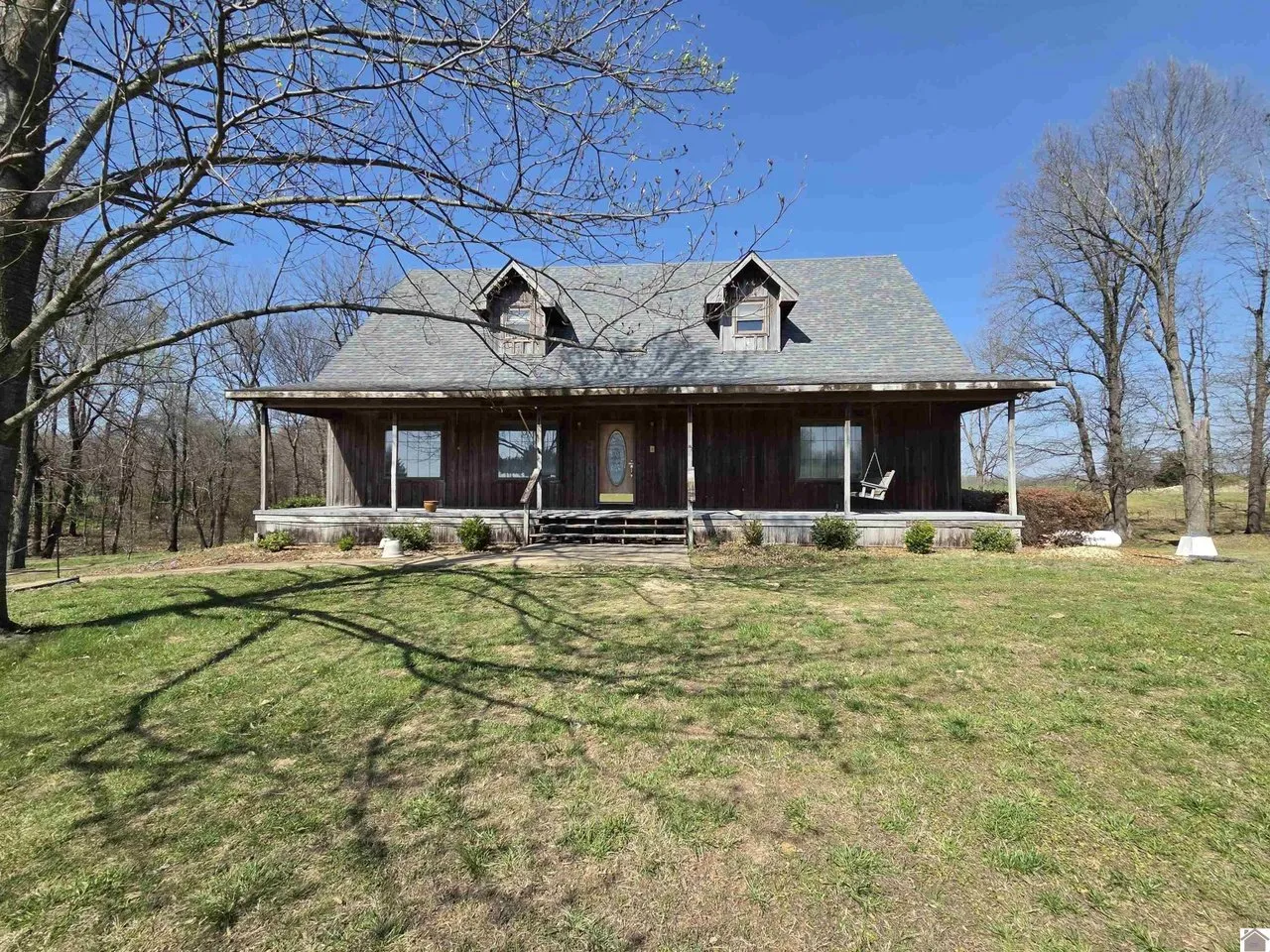 A dark two-story house with dormers and a full-width porch on a mown lawn under bare trees, near Clinton, Kentucky